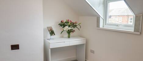 A small, white room with a slanted ceiling, a white desk, a vase with flowers, and a framed picture on the desk.
