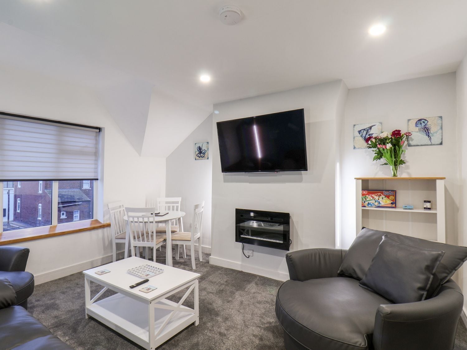 Una sala de estar moderna con un televisor de pantalla plana, un comedor con sillas blancas, un sofá gris y una estantería con un jarrón de flores.