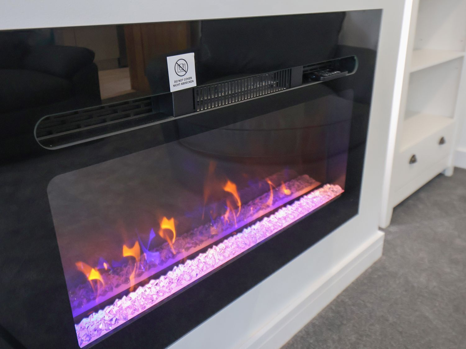 A fireplace with a glass enclosure, a 'DO NOT COVER' sign, and a white cabinet to the right.