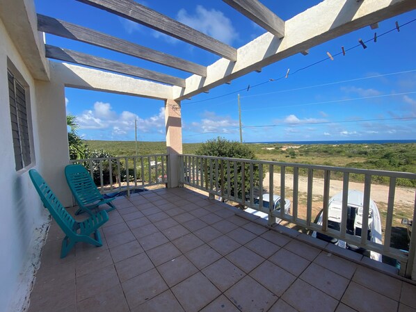 A covered balcony with tiled flooring, two green plastic chairs, and a view of the ocean and greenery.