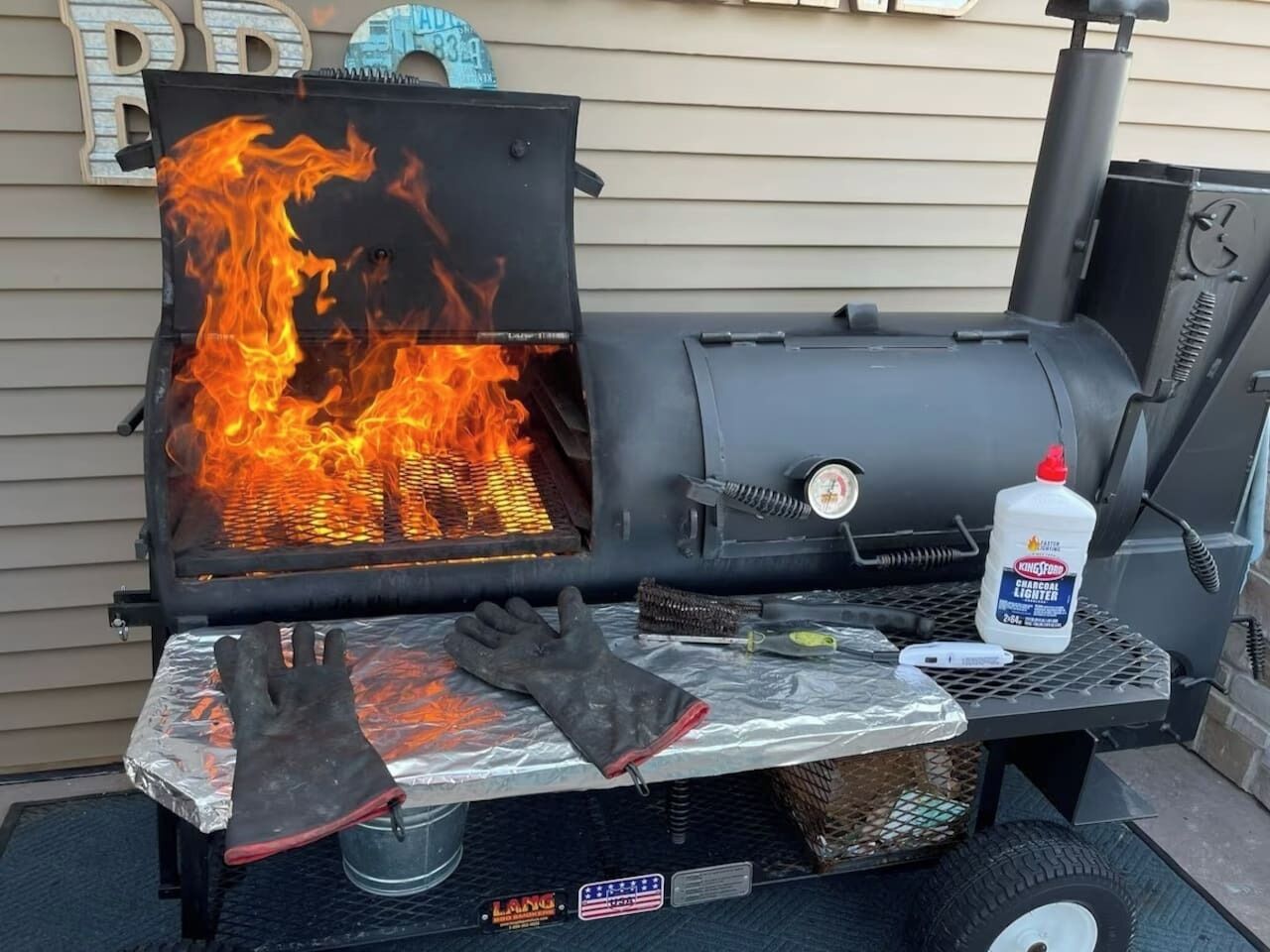 A barbecue grill with flames, a pair of gloves, a lighter, and a grill brush on a metal table.