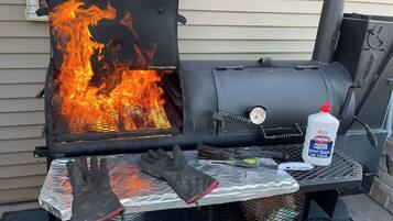 A barbecue grill with flames, a pair of gloves, a lighter, and a grill brush on a metal table.