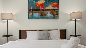 A hotel room with a bed, two bedside lamps, a cityscape painting, and a wooden headboard. - Steps to SoCo, Mins to Zilker | Quiet Pet-Friendly Casita w Grill + Workspace (Austin)