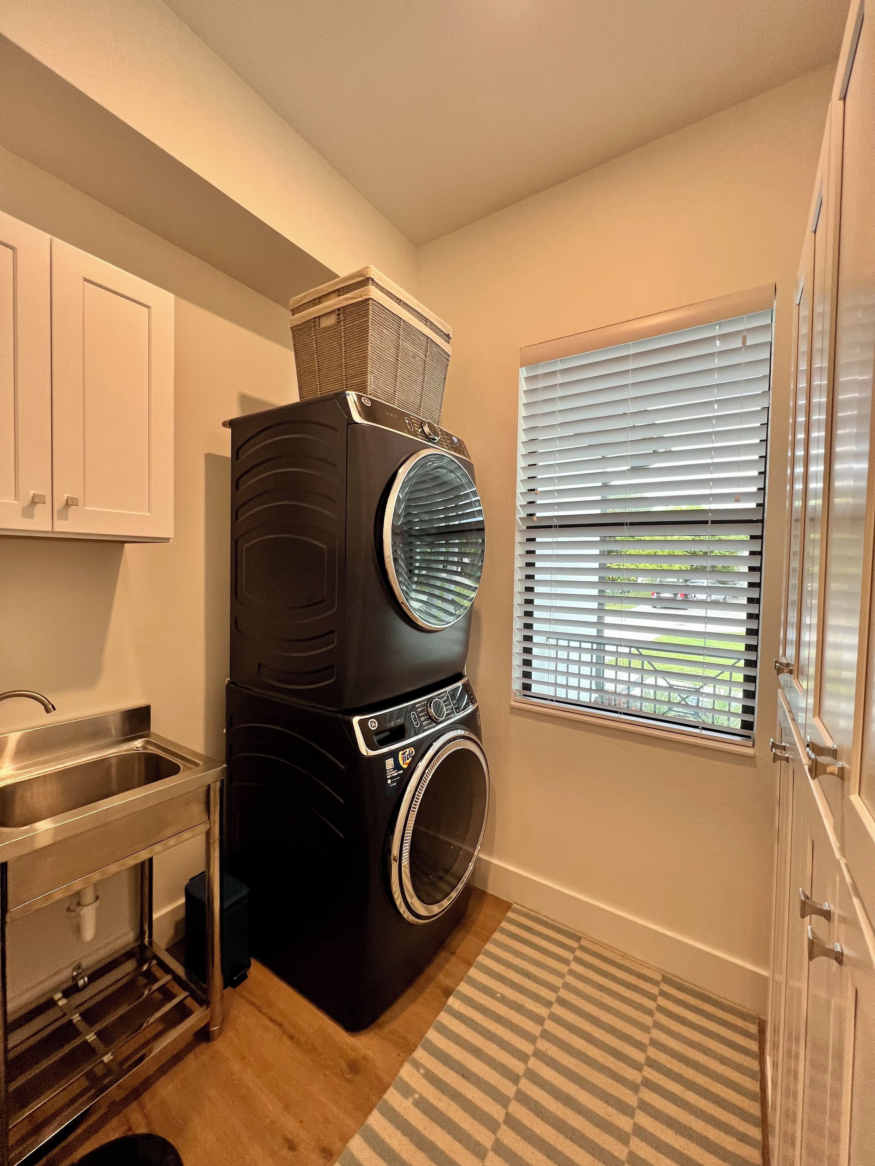 A compact laundry room with a front-loading washer and dryer, a stainless steel sink, and a window with blinds.