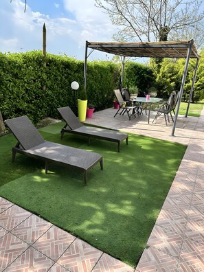 A patio with artificial grass, two lounge chairs, a table with chairs, and a pergola.