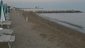 A beach with rows of sun loungers, a rocky breakwater, and a clear sky.