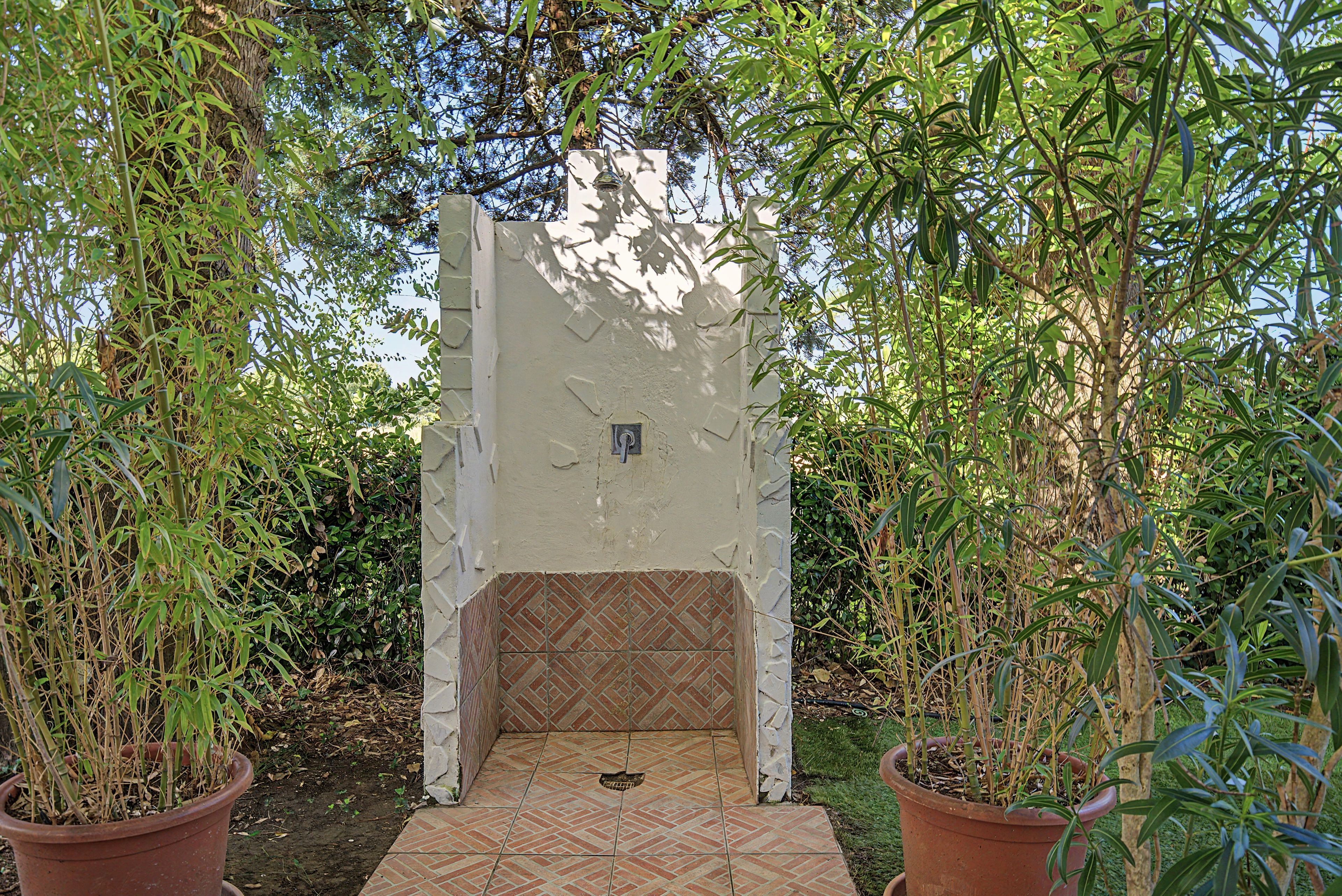 A covered outdoor area with a tiled floor, surrounded by potted plants and trees.