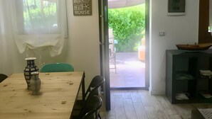 Ein Essbereich mit Holztisch, Stühlen und einer Vase. Eine Tür führt nach draußen, ein kleines Regal mit einem Bootsmodell und eine Wand-Klimaanlage sind ebenfalls vorhanden.