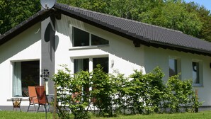 Een twee verdiepingen tellend huis met een witte gevel, een donker dak en een schoorsteen. Er is een raam met gordijn, een deur en een klein balkon met een stoel en een tafel.