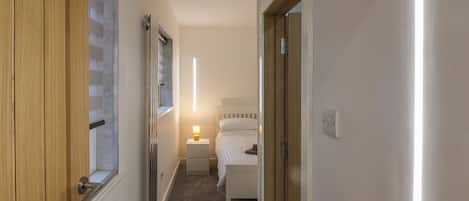 Un pasillo estrecho que conduce a un dormitorio con cama, mesita de noche con lámpara y ventana.