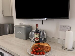 A kitchen counter with a loaf of bread, a bottle of wine, two glasses, a plate with tomatoes, a bread box, and a TV. - Frankies Seaview 3 (Bridlington)