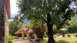 Un espace extérieur abrité, avec des tables et des chaises, un bâtiment en pierre et un grand arbre.