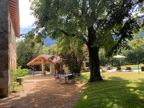 Un espace extérieur abrité, avec des tables et des chaises, un bùtiment en pierre et un grand arbre.