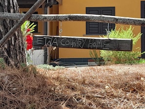 A rustic wooden sign with "EIA COWRY HOME" written on it, set against a yellow building with black shutters.