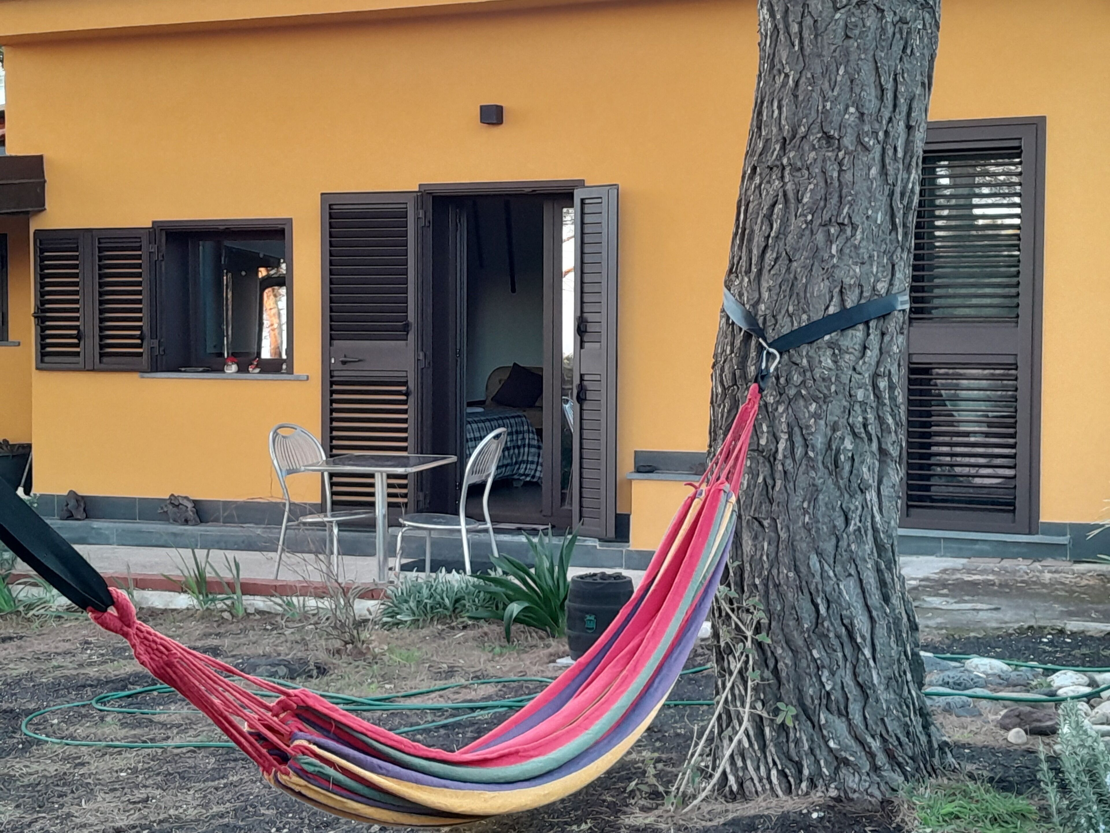 Un'amaca legata a un albero, un tavolino con sedie e un edificio con pareti gialle e persiane scure.