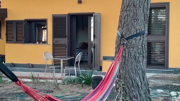 Un'amaca legata a un albero, un tavolino con sedie e un edificio con pareti gialle e persiane scure.