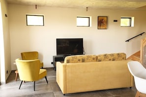 Een woonkamer met een bank, een gele fauteuil, een televisie en een trap.