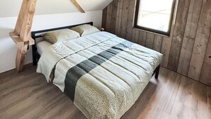 Makuuhuone, jossa on kalteva katto, puupalkit, kuviollinen päiväpeite ja ikkuna.
