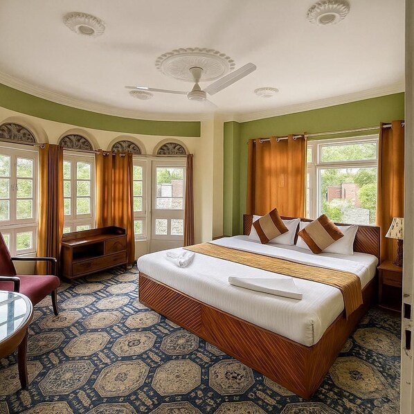 Quarto de hotel com uma cama grande, uma escrivaninha, uma cadeira, um ventilador de teto e vista para o verde lá fora.