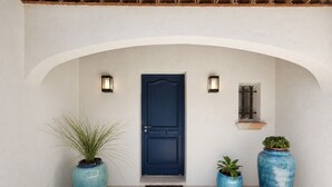 Une porte bleue, deux lampes fixées au mur de part et d’autre, une petite fenêtre et trois grands pots de plantes.