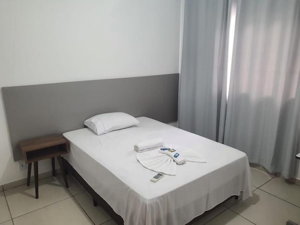 A hotel room with a bed, a nightstand, a remote control, and a window with curtains.