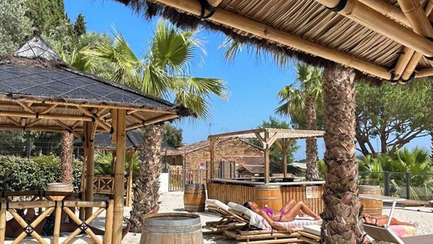 A beachside resort with wooden structures, palm trees, and a sandy area with lounge chairs. - Mobil home for 6 guests – Gulf of Saint-Tropez 4★ (La Garde-Freinet)
