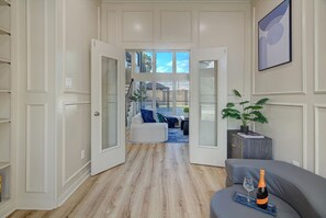 Ein geräumiges Zimmer mit großem Fenster, einer Couch, einem kleinen Tisch mit Flasche und Glas sowie einer Pflanze.