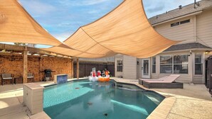 Una piscina con cubierta de madera, sillas de descanso y un área sombreada con grandes toldos.