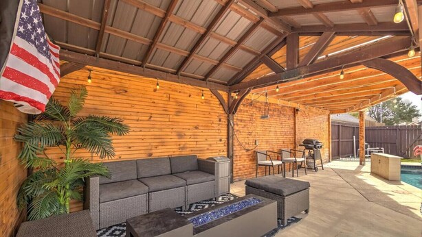 Un patio exterior con terraza de madera, un sofá gris, un brasero y una piscina al fondo.