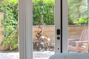A view through a glass door of a patio area with a brick wall, a wooden chair, and a plant with red and yellow leaves. - South Mission Sand & Surf Retreat with AC & 3 Car Garage! (San Diego)