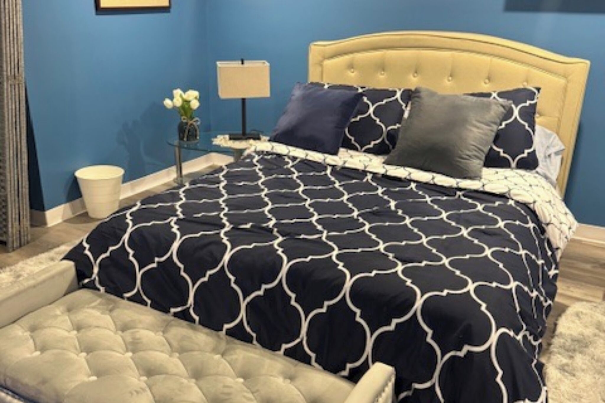 A bedroom with a bed, a nightstand, a lamp, a vase with flowers, and a chair.