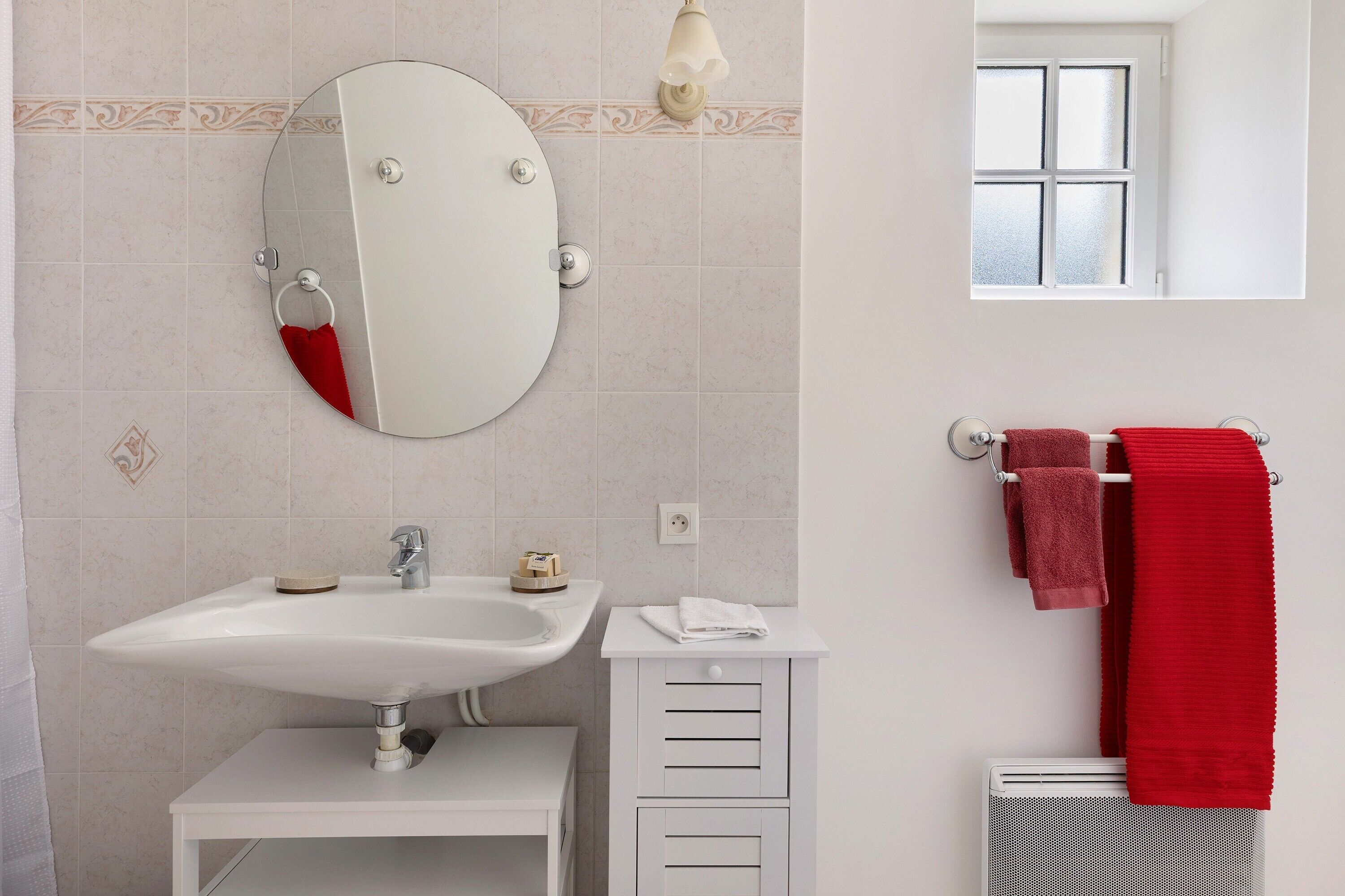 Une salle de bain avec un miroir rond, un lavabo blanc et un porte-serviettes avec des serviettes rouges.