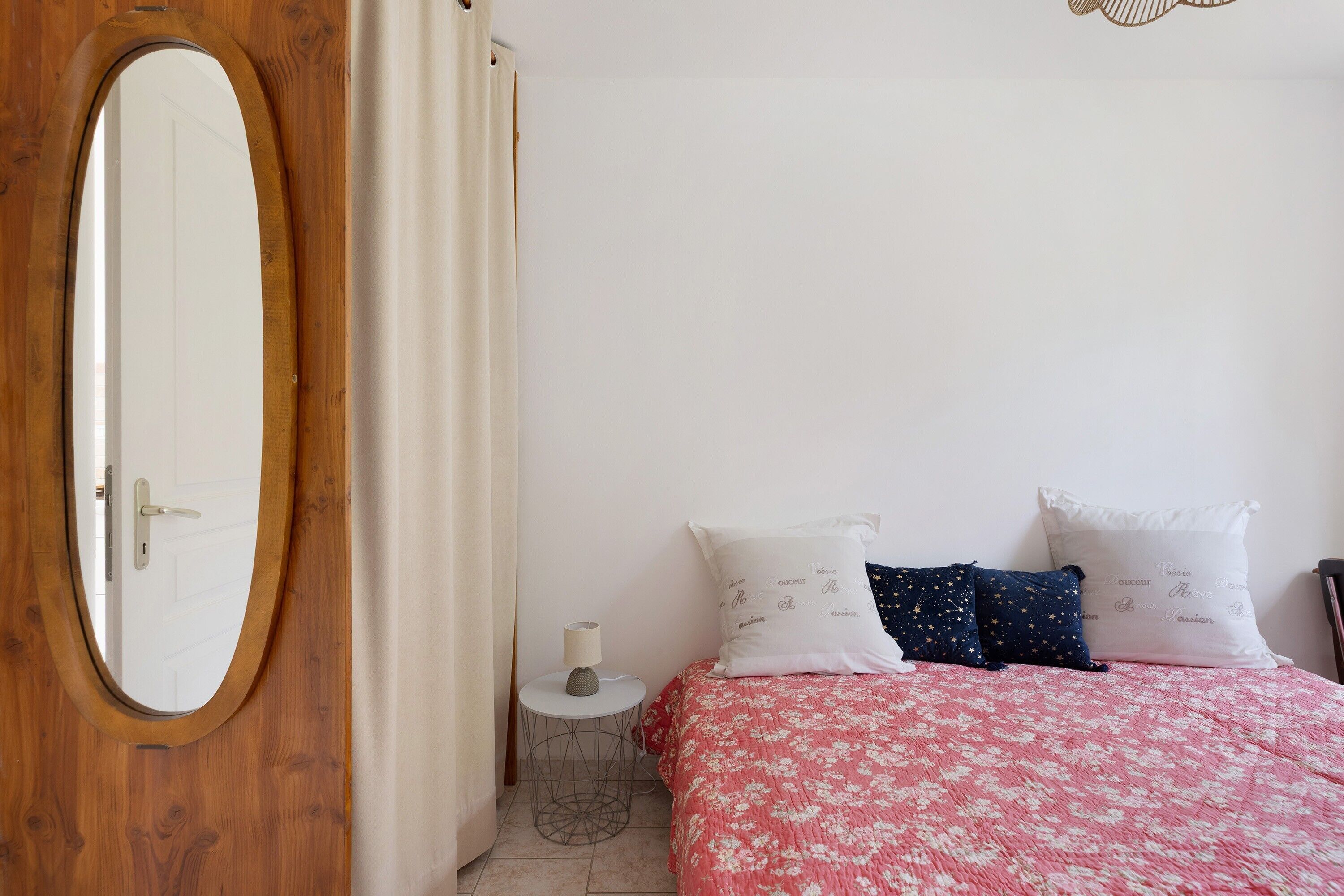 Een slaapkamer met een houten deur, een ronde spiegel, een klein rond tafeltje met een lamp, een bed met een bloemenbedekking en twee kussens met tekst.