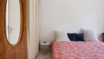 Een slaapkamer met een houten deur, een ronde spiegel, een klein rond tafeltje met een lamp, een bed met een bloemenbedekking en twee kussens met tekst.