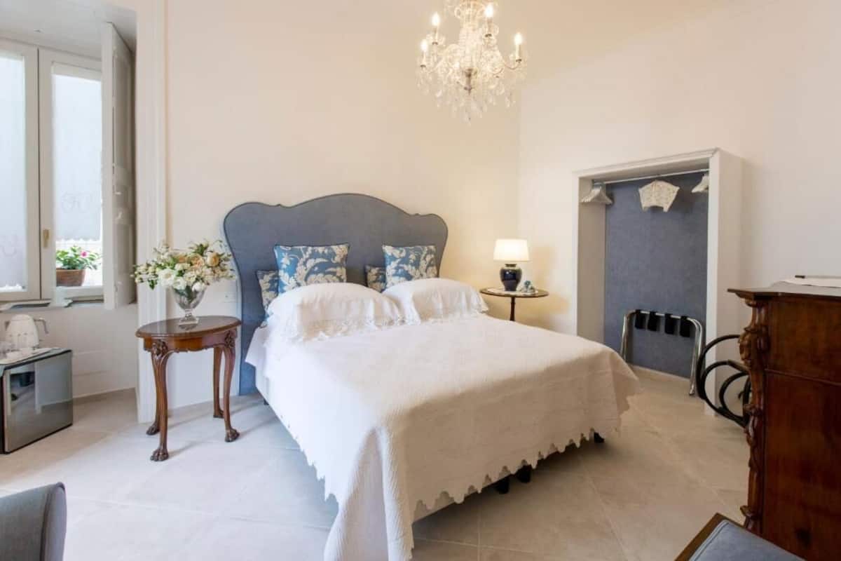 A bedroom with a bed, a chandelier, a nightstand, a dresser, and a window with a view.