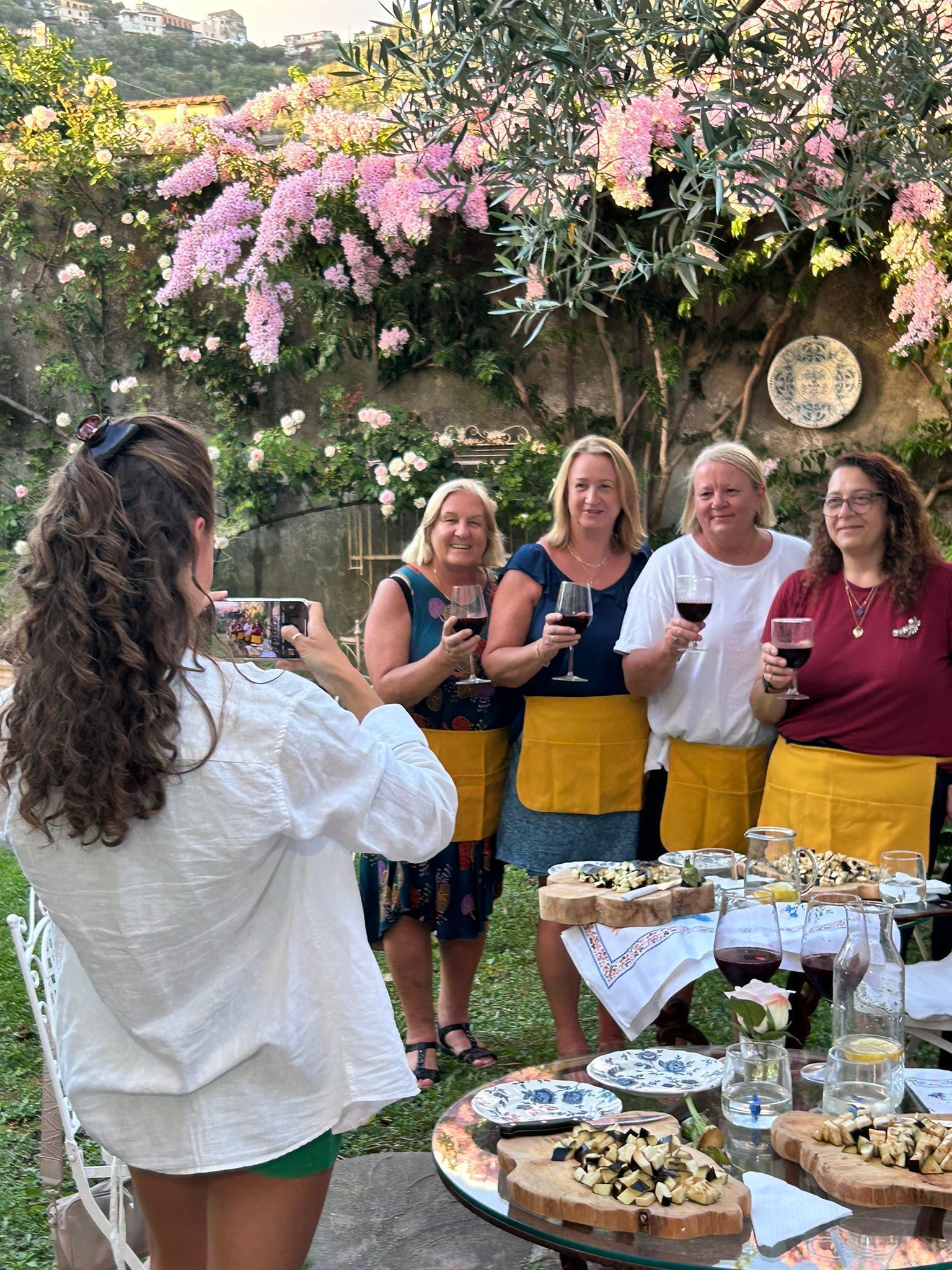 Un groupe de personnes s’est réuni autour d’une table où étaient disposés de la nourriture et des boissons, dans un cadre extérieur agrémenté de verdure et de fleurs.