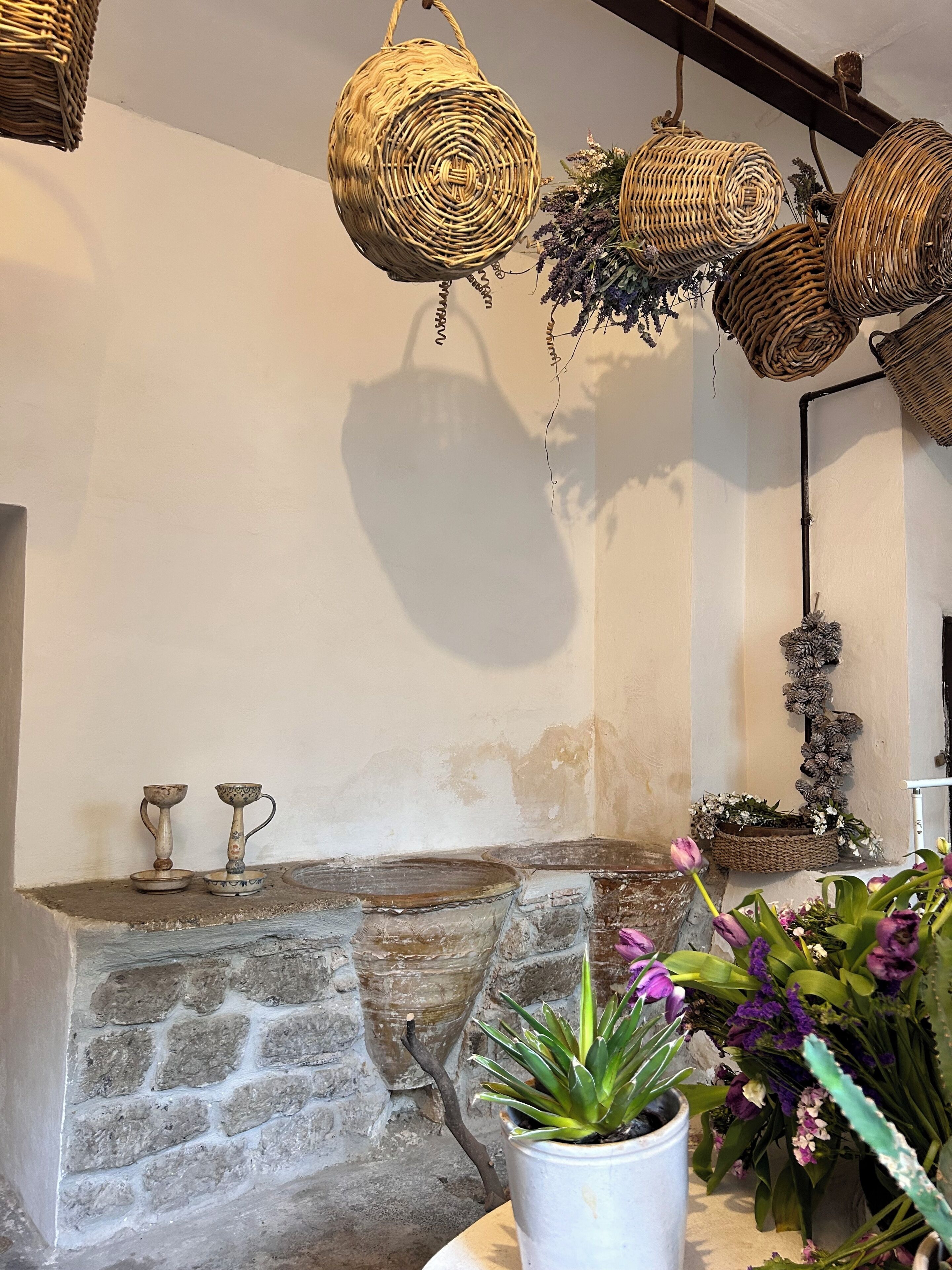 Uma lareira de pedra com duas taças decorativas, cestos de vime pendurados acima e um vaso com flores roxas.