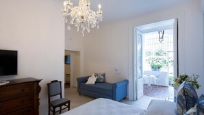 A bedroom with a bed, dresser, TV, chandelier, and a view to an outdoor patio. - Palazzo Cacace Relais (Meta)