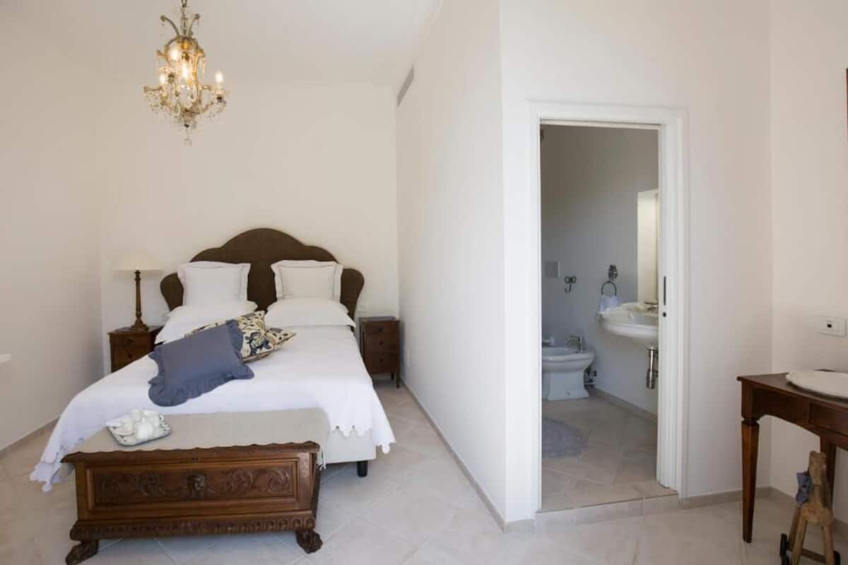 A bedroom with a wooden bed, a nightstand, a bedside table, a chandelier, and a bathroom visible through an open door.