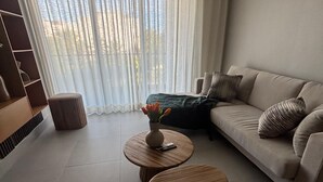A modern living room with a sofa, two round wooden tables, a vase with flowers, and a large window with sheer curtains. - Beautiful 1-Bedroom Apt en las Canas punta cana. (Punta Cana)