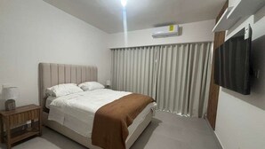 A bedroom with a bed, a bedside table, a wall-mounted air conditioner, and a curtain. - Beautiful 1-Bedroom Apt en las Canas punta cana. (Punta Cana)