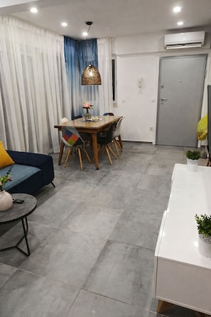 Una sala de estar moderna con un sofá, una mesa de comedor y sillas. Hay una mesita con un florero y un control remoto. La habitación tiene un ventanal con cortinas, un equipo de aire acondicionado y una puerta.
