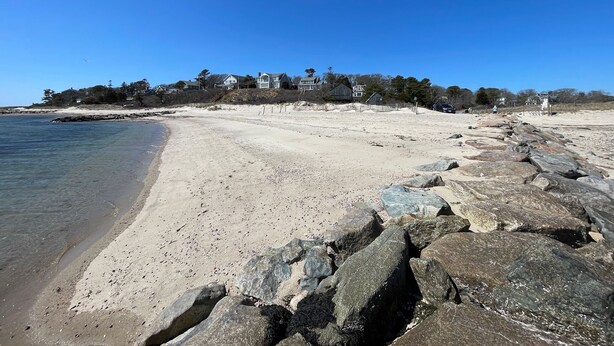 Ein Strand mit felsigem Ufer, blauem Himmel und Häusern im Hintergrund.