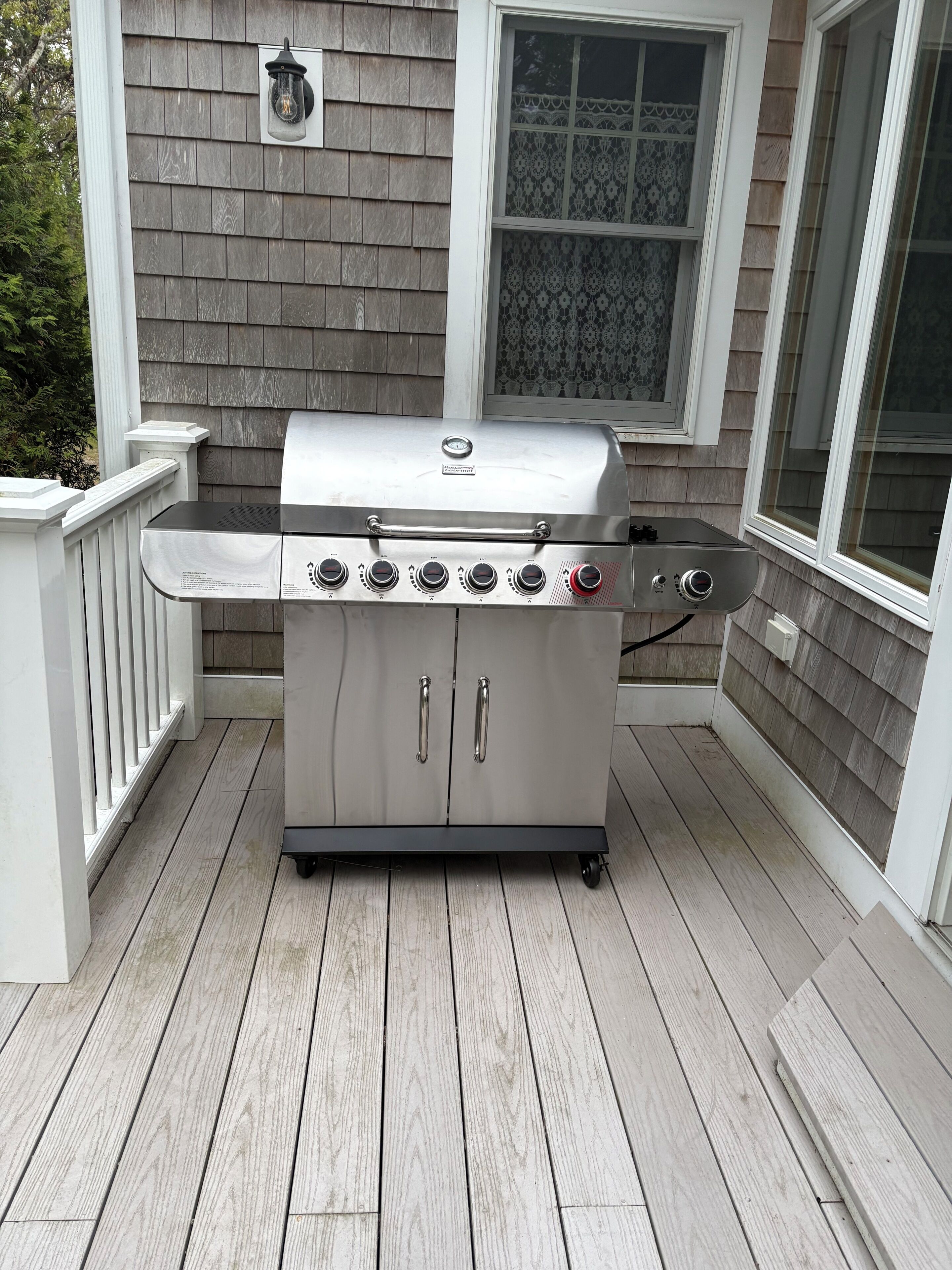 A stainless steel barbecue grill with four burners and a side burner, positioned on a wooden deck with a white railing and a window with patterned glass.