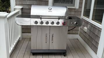 A stainless steel barbecue grill with four burners and a side burner, positioned on a wooden deck with a white railing and a window with patterned glass.