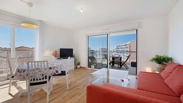 Ein modernes Wohnzimmer mit einer Couch, einem Esstisch, Stühlen, einem Fernseher und einem Balkon mit Blick auf Gebäude.