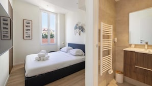 Ein Schlafzimmer mit Bett, Nachttisch, Fenster und einem Badezimmer mit Waschbecken und Spiegel.