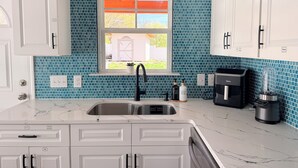 Een keuken met witte kasten, een blauwe betegelde spatwand, een raam met uitzicht, een spoelbak en een koffiezetapparaat.