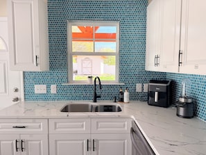 A kitchen with white cabinets, a blue-tiled backsplash, a window with a view, a sink, and a coffee maker.