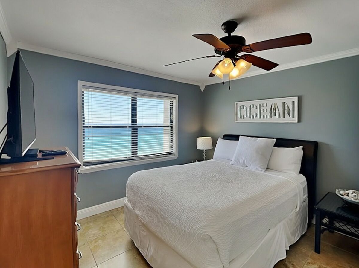 A bedroom with a bed, ceiling fan, window, dresser, and a wall-mounted lamp.